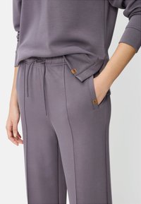 Lyse lilla afslapningssæt bestående af en løs top og bukser med snøre og sidelommer. Stoffet har en glat tekstur; logoaccenter er synlige.