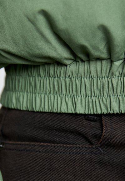 Zielona elastyczna talia z materiału, z teksturowanymi fałdami, połączona z czarnym dżinsowym materiałem, prezentującym gładkie wykończenie i kontrast kolorystyczny.