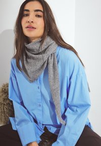 Harmaa villahuivi kaulan ympärillä, yhdistettynä vaaleansiniseen nappipaitaan. Pehmeä rakenne ja rento istuvuus lisäävät asun mukavuutta.