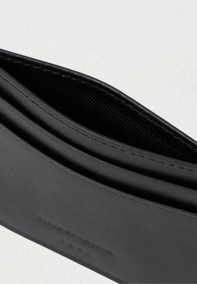 Carteira de couro preta parcialmente aberta, mostrando o exterior liso e as bordas cosidas contra um fundo claro e liso.