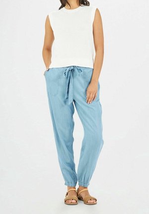 Nainen pukeutuneena valkoiseen hihattomaan neulepaitaan, vaaleansinisiin vetoköysillä varustettuihin housuihin, joissa on joustavat lahkeet, ja ruskeisiin punottuihin remmisandaaleihin, seisoo käsi taskussa.