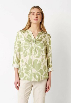 Woman wearing a light green and cream floral buttoned blouse with rolled-up sleeves and light beige pants against a plain background.