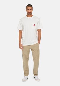 Weißes Baumwoll-T-Shirt mit einem roten Grafikdesign auf der linken Brusttasche, kombiniert mit beigen Kordhosen und weißen Sneakers.