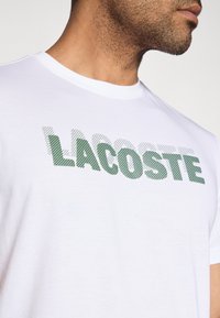 Biała bawełniana koszulka z napisem „LACOSTE” w zielone kropki, okrągły dekolt, krótkie rękawy i gładka tekstura.