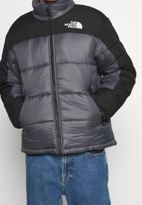 Vatteret jakke i sort og grå med høj krave, fuld lynlås og logo på brystet; har quiltede sektioner og sidelommer.