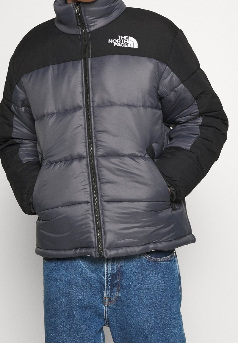 Vatteret jakke i sort og grå med høj krave, fuld lynlås og logo på brystet; har quiltede sektioner og sidelommer.