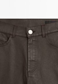 Detail tmavě hnědého pasu kalhot s knoflíkovým zapínáním, poutky na opasek a viditelnými předními kapsami a šicími detaily.