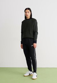 Tamnozeleni rebrasti pulover preko crne majice s dugim rukavima, u kombinaciji sa crnim trenirkama s bijelim bočnim prugama i bijelim tenisicama.