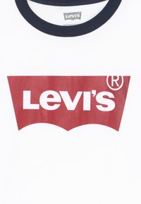 Camiseta blanca con un atrevido logo rojo de Levi's en forma de ala de murciélago. Cuello redondo en color azul marino y material de algodón suave. Diseño sencillo.