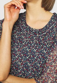 Blouse florale avec un fond bleu marine, ornée de petites fleurs rouges et crème, un col rond et des manches courtes bouffantes avec un détail froncé.