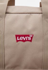 Sac en tissu beige avec une surface texturée, présentant un logo Levi's brodé rouge et blanc bien en évidence à l'avant.