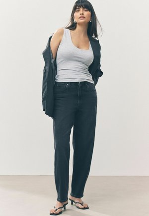 Grijze tanktop, zwarte high-waisted jeans en zwarte hakken. Model draagt een zwart blazer, met steil donker haar en minimale accessoires.