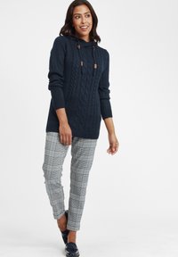 Marinblå stickad tröja med kabeldesign och huva, matchad med rutiga byxor med blå och svarta detaljer. Platta skor fulländar outfiten.