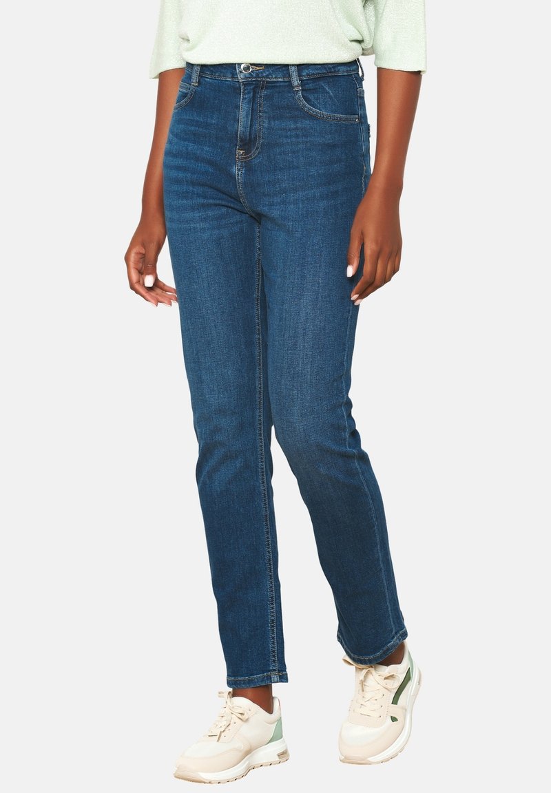 Tamsiai mėlynos aukšto liemens džinsai su tiesiomis kojomis, turintys priekinės kišenes ir subtilius siuvinėjimo akcentus, derinami su baltai žaliais sportbačiais.