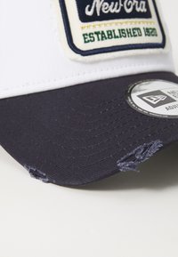Gorra blanca y azul marino con un visera texturizada, que cuenta con un parche cosido etiquetado "New Era Established 1920" y un borde desgastado.