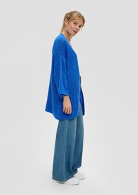 Oversize blauwe gebreide cardigan met een losse pasvorm, geribbelde textuur en brede mouwen, gecombineerd met high-waisted denim broeken en witte sneakers.