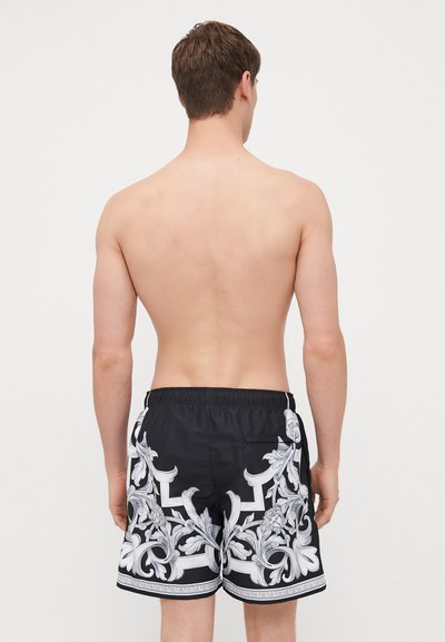 Shorts de bain noirs avec des motifs floraux blancs ornés, taille élastique et une poche arrière. Vue de dos.