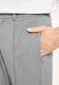 Pantalones grises con una textura sutil, que cuentan con un bolsillo lateral. Una mano reposa sobre el bolsillo, destacando su diseño práctico.