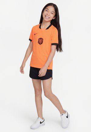Fille souriante en maillot de sport orange et short noir, debout avec un pied légèrement en avant sur un fond blanc uni.