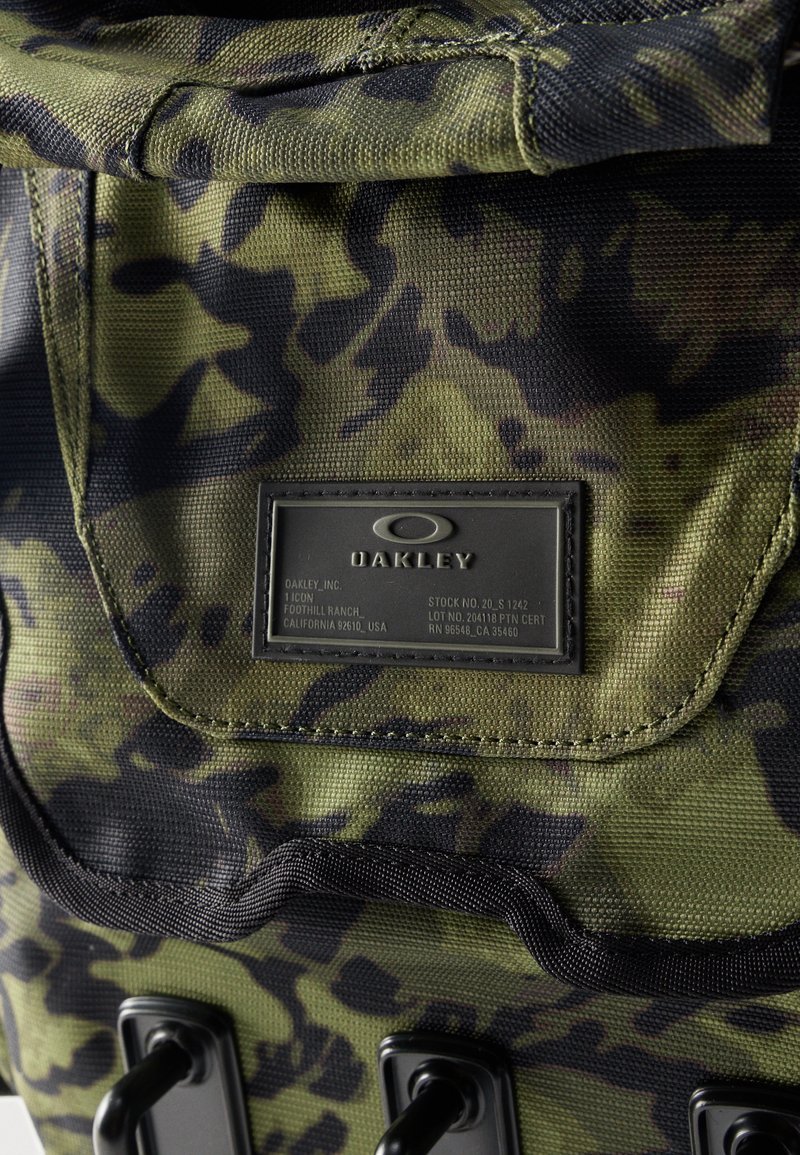 Grønt og sort camouflage-stof med et Oakley-mærke syet på en rygsæk-lomme og to sorte plastikspænder nedenunder.
