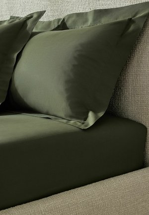 Nahaufnahme von olivgrünen Kissenbezügen und einem Spannbettlaken auf einem beige strukturierten Bettrahmen.