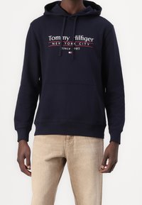 Felpa blu navy con tasca frontale a marsupio, coulisse regolabile e logo con "Tommy Hilfiger, New York City" in testo bianco e rosso.