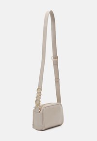 Borsa crossbody in finta pelle beige con chiusura a zip, forma rettangolare, tracolla regolabile e dettagli circolari dorati sulla tracolla.
