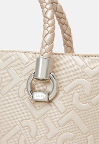 Sac à main beige en matériau texturé, avec une poignée tressée, des anses en métal argenté et un logo en relief.