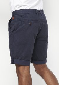 Man in marineblauwe opgerolde shorts met zij- en achterzakken, gecombineerd met een witte ingestopte shirt, gezien vanaf de achterkant van de taille naar beneden.