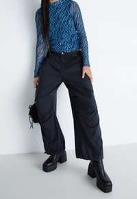 Blå och svart långärmad topp med ett zebramönster, i kombination med vida svarta byxor och kraftiga svarta klackstövlar, hållandes en liten väska.