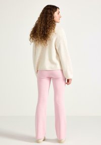 Licht beige oversized trui met lange mouwen, gecombineerd met nauwsluitende roze flarebroeken. Model met krullend haar, gekeerd van de camera vandaan.
