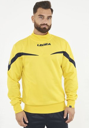 Uomo con la barba che indossa una felpa LEGEA gialla con dettagli a strisce nere, in piedi davanti a uno sfondo bianco uniforme.