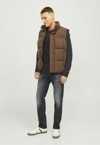 Bruine puffer vest over een donkere trui, donkere jeans en witte sneakers. De vest heeft een hoge kraag en een gewatteerd ontwerp.