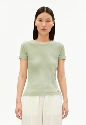 Jonge vrouw met steil donker haar, gekleed in een lichtgroen nauwsluitend topje met korte mouwen en losse witte broek, tegen een witte achtergrond.