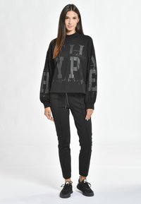 Hanorac negru supradimensionat cu litere grafice, asociat cu pantaloni de jogging negri ajustați. Materialele par moi, având un stil casual sport.