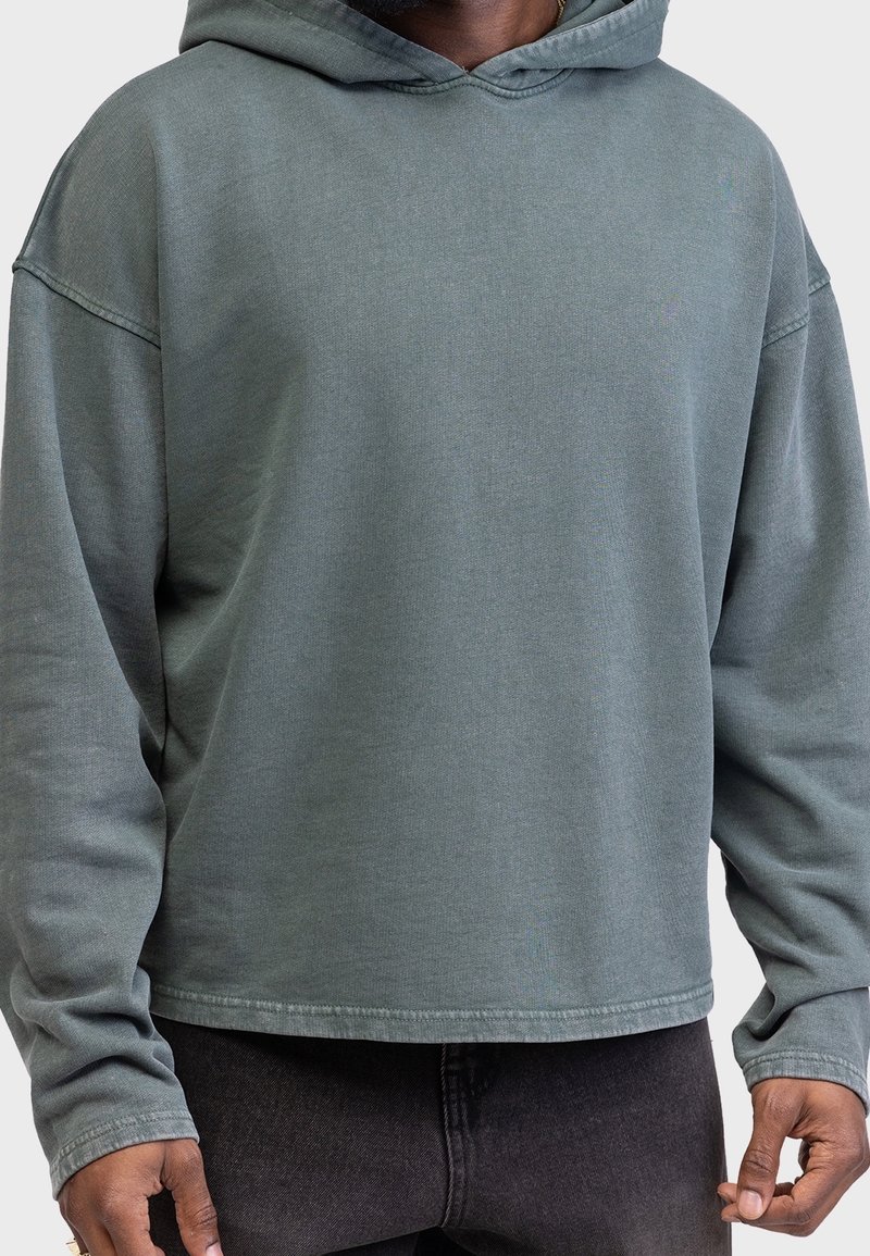 Personne portant un sweat à capuche vert délavé et ample ainsi qu’un jean sombre, debout, les bras détendus le long du corps, devant un fond uni.