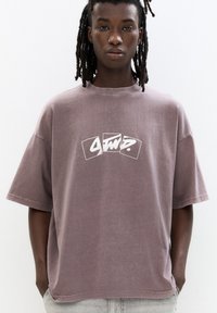 Større, kortærmet t-shirt i dæmpet mauve med et hvidt grafisk logo trykt på fronten. Fremstillet af bomuld med en blød struktur.