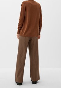 Hnědý pletený svetr s dlouhými rukávy v kombinaci se širokými kalhotami hnědého vzoru psí zub. Černé baleríny, jednobílé pozadí.