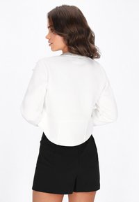 Valkoinen lyhyt takki, jossa pitkät hihat, koristeltu hopeisella kauluksella ja sileällä pinnalla, yhdistetty mustiin shortseihin.