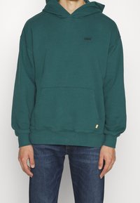 Zielona bluza z kapturem uszyta z miękkiego materiału, z kieszenią kangurką, ściągaczami przy rękawach i małym logo na klatce piersiowej. W połączeniu z dżinsami.