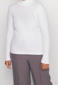 Haut à col roulé blanc à manches longues fabriqué dans un matériau lisse et extensible ; présente un petit logo sur la poitrine. Coupe ajustée avec des manches fines.