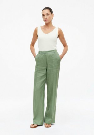 Vrouw draagt een wit mouwloos topje, losse lichtgroene broek en sandalen, staat met handen in de zakken tegen een effen witte achtergrond.