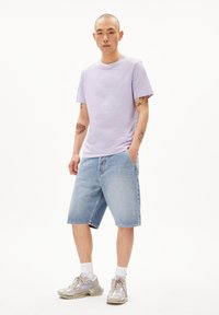 Helles lila Baumwoll-T-Shirt mit kurzen Ärmeln; hellblaue Jeans-Shorts; graue Sneakers mit lila Akzenten; Tattoos sichtbar auf den Armen.