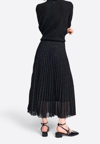 Jupe plissée noire avec des paillettes métalliques, superposition transparente et taille élastique. Associée à des chaussures noires à talons bas et à lanières.