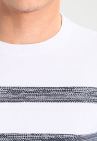 Gros plan sur le cou et le haut de la poitrine d'une personne portant un pull blanc avec deux rayures horizontales tricotées en noir et gris.