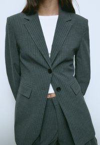 Siv blazer s črtami in dvema sprednjima žepoma ter črnimi gumbi, v kombinaciji z ujemajočimi hlačami. Tkanina ima gladko teksturo.