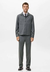 Veste de costume gris texturé avec deux poches avant, associée à un pantalon slim assorti. Portée avec une chemise blanche et une cravate sombre, ajoutée avec des chaussures noires.
