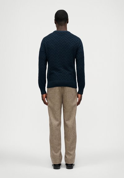 Dunkelblauer, strukturierter Strickpullover mit Rundhalsausschnitt, kombiniert mit hellbraunen Tweedhosen und schwarzen Schuhen, von hinten betrachtet.