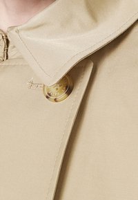 Beige kappa med bred krage, dragkedjestängning och en framträdande rund knapp med "RALPH LAUREN"-logotyp. Slät tygtextur.