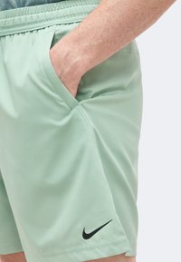 Το χέρι ενός ατόμου στην τσέπη ενός ανοιχτοπράσινου σορτς Nike με μαύρο λογότυπο swoosh στο αριστερό πόδι, σε ουδέτερο φόντο.